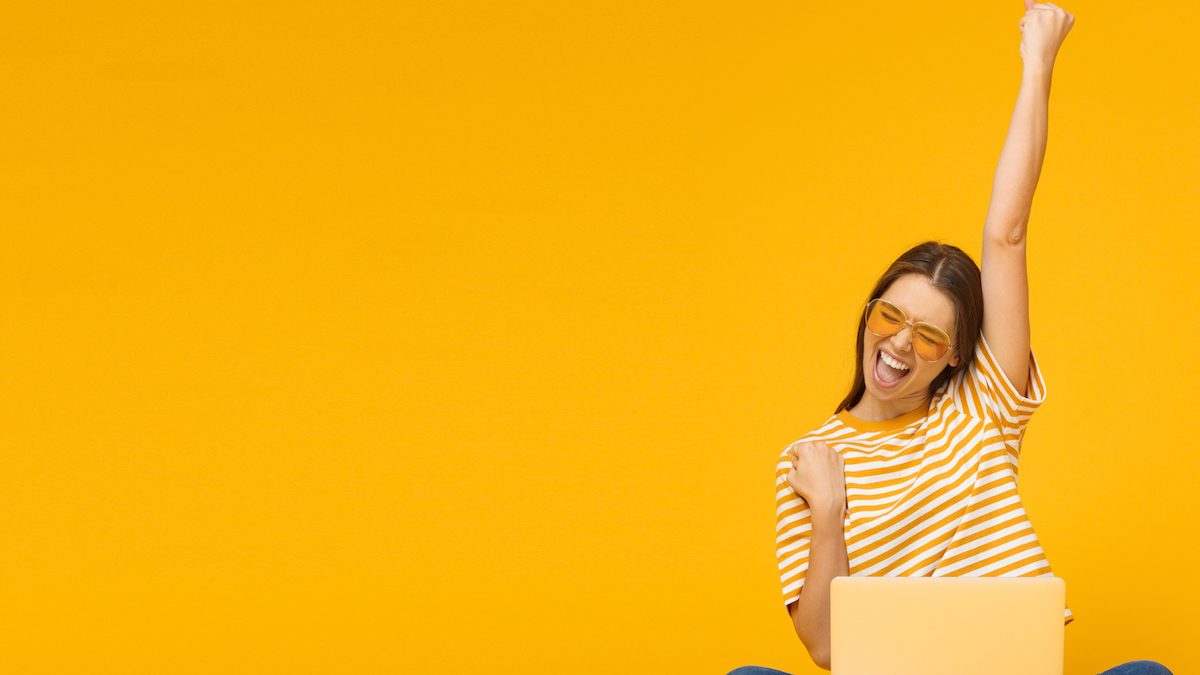 Winner! Excited smiling girl sitting on floor with laptop, raising one hand in the air is she wins, isolated on yellow background (Winner! Excited smiling girl sitting on floor with laptop, raising one hand in the air is she wins, isolated on yellow b